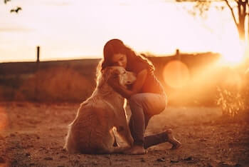 A woman shown kneeling down and hugging her Golden Retriever. There is a sunset in the background.